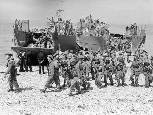 Photographie en noir et blanc – De nombreux soldats en tenue de combat marchent sur une plage rocheuse à partir des rampes d’une péniche de débarquement. Des membres de la marine attendent le débarquement d’une deuxième péniche.